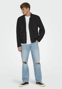 Svart mockajacka med fickor, bärs över en vit t-shirt, i kombination med ljusblå slitna jeans och svarta sneakers.