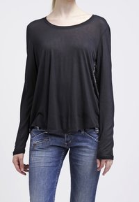 Zwarte blouse met lange mouwen, een losse pasvorm, ronde halslijn en transparante textuur. Gecombineerd met blauwe jeans met ritszakken.