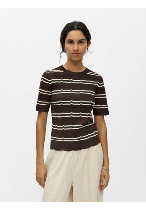 Mujer con cabello oscuro que lleva una camiseta de punto marrón de manga corta con rayas horizontales blancas y pantalones anchos de color beige claro, de pie frente a un fondo blanco.