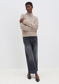 Helle beige Strickjacke mit hohem Kragen, kombiniert mit lockeren, dunkelgrauen Jeans und braunen spitzen Schuhen. Strukturierter Stoff.