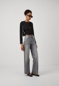 Donna che indossa un crop top nero con lacci, occhiali da sole oversize neri, jeans grigi a gamba larga, ballerine nere con borchie e una borsa a spalla nera in piedi.