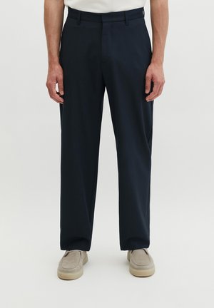 Mann trägt schwarze maßgeschneiderte Hose und beige Slipper, steht vor einem schlichten hellen Hintergrund.