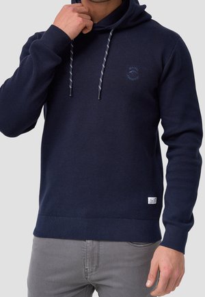 Mann trägt eine marineblaue Kapuzenjacke mit gestreiften Kordeln und kleinem Logo auf der Brust, kombiniert mit grauer Hose vor einem einfarbigen Hintergrund.