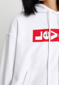 Felpa bianca con cappuccio decorata da un grande logo rettangolare rosso con la scritta "Levi's". Presenta una tasca frontale e un cappuccio regolabile con cordini. Tessuto morbido.