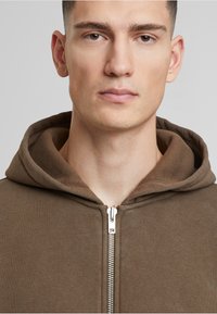 Brauner Reißverschluss-Hoodie mit weicher Textur, ausgestattet mit einer Kapuze mit Kordelzug und einem metallischen Reißverschluss. Nahaufnahme betont den Kragen und die Stoffdetails.