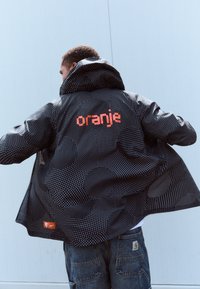 Crna jakna s uzorkom bijelih točkica, veliki narančasti natpis "oranje" na leđima i mali Nike logo na donjoj lijevoj strani. Ima kapuljaču.