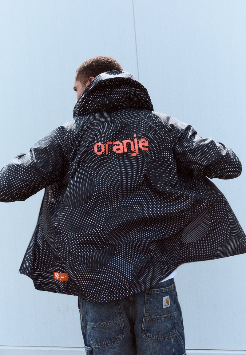 Crna jakna s uzorkom bijelih točkica, veliki narančasti natpis "oranje" na leđima i mali Nike logo na donjoj lijevoj strani. Ima kapuljaču.