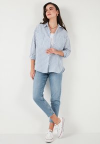 Blau-weiß gestreiftes Button-Up-Hemd, lange Ärmel, zwei Fronttaschen, über einem weißen Tanktop getragen, kombiniert mit hellblauen Jeans und weißen Turnschuhen.