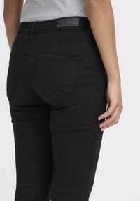 Femme portant un jean noir ajusté et un haut gris côtelé, montrée de la taille aux pieds, mettant en valeur les poches arrière et l'étiquette à la taille.