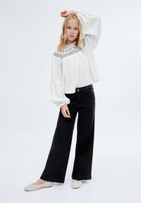 Vit långärmad blus med vertikala ränder och broderad geometrisk halsringning, tillsammans med svarta vida jeans och ljusa skor.