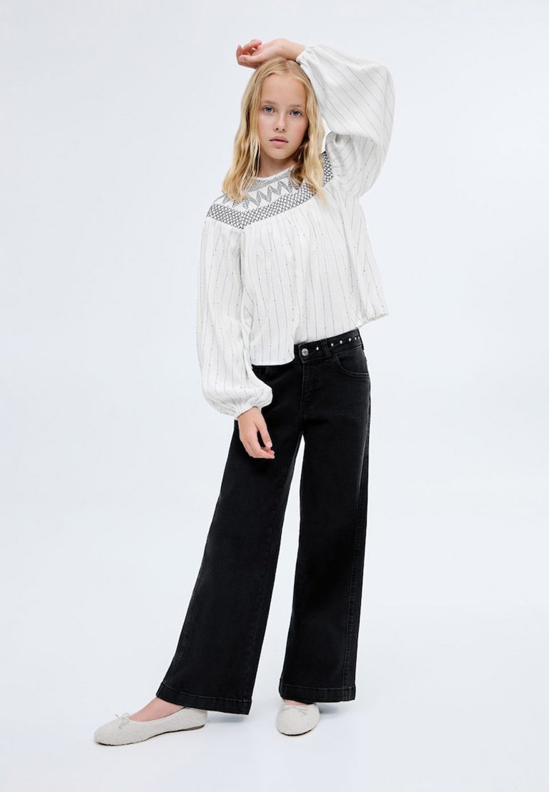 Vit långärmad blus med vertikala ränder och broderad geometrisk halsringning, tillsammans med svarta vida jeans och ljusa skor.