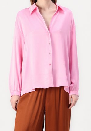 Kobieta ubrana w luźną, różową koszulę na guziki z długim rękawem, w połączeniu z szerokimi, brązowymi spodniami na nogawki, na białym tle.