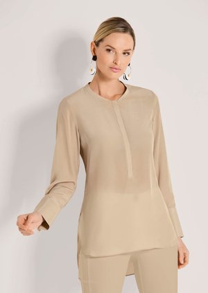 Frau trägt eine beige langärmelige Tunika und Hose, accessoriert mit großen runden schwarz-weißen Ohrringen, posiert vor einfachem Hintergrund.