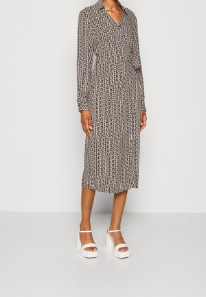 Femme portant une robe portefeuille mi-longue avec un motif géométrique noir et blanc et des sandales à plateforme blanches à bouts ouverts, debout devant un fond uni.