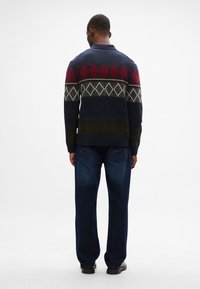 Sweter dzianinowy w ciemnoniebieskim kolorze, z diamentowym wzorem w czerwieni, zieleni i kremie. Połączony z ciemnoniebieskimi dżinsami i czarnymi butami.