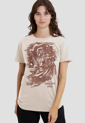 Woman wearing beige t-shirt with brown graphic of Mandalorian holding Grogu and smoking weapon, text "Mandalorian and Grogu."