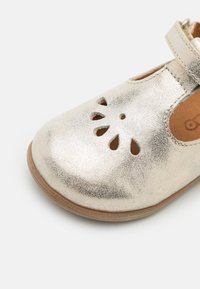 Zapato de cuero plateado brillante con diseño floral recortado en la puntera, cierre de correa y suela de goma beige. Textura suave y forma redondeada.