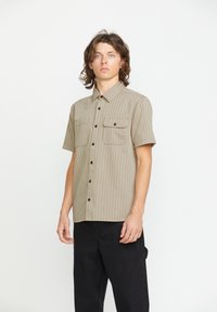 Kortærmet, knaplukket skjorte i beige med lodrette striber, to brystlommer og kontrasterende sorte knapper. Stoffet ser tekstureret ud.