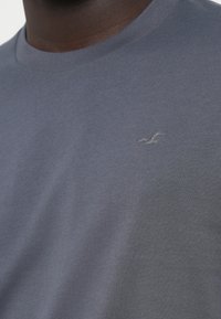 Tricou din bumbac gri cu guler rotund, având un mic logo tonal sub formă de pasăre pe partea stângă a pieptului și o textură netedă și uniformă.