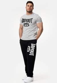 Szürke pamut póló, amelyen vastag fekete betűkkel szerepel a "TAPOUT" felirat, fekete melegítőnadrággal párosítva, amelyen fehér "TAPOUT" minták láthatók, fehér sportcipővel viselve.