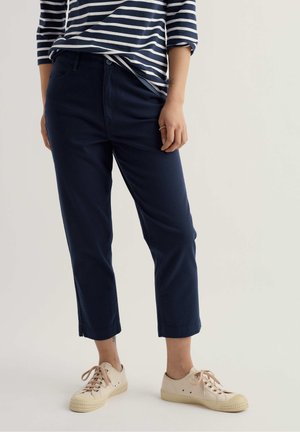 Person wearing navy cropped pants, striped long-sleeve shirt tucked in, and beige lace-up sneakers standing against a plain background.