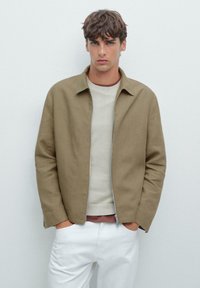Giacca leggera marrone con colletto, chiusura a zip e due tasche laterali, indossata sopra un maglione crema e pantaloni bianchi. Tessuto liscio.