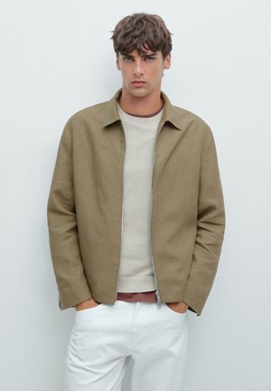 Veste légère marron avec un col, fermeture éclair à l'avant et deux poches latérales, portée par-dessus un pull crème et un pantalon blanc. Texture lisse.
