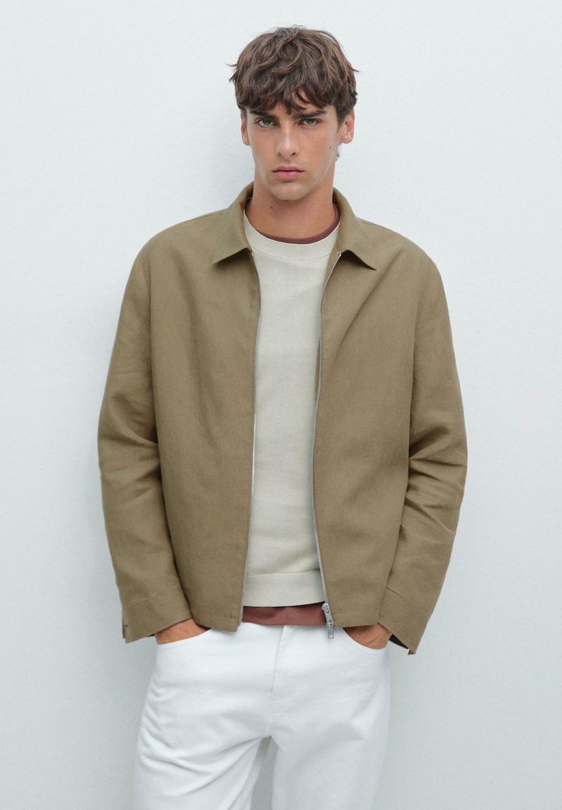 Giacca leggera marrone con colletto, chiusura a zip e due tasche laterali, indossata sopra un maglione crema e pantaloni bianchi. Tessuto liscio.