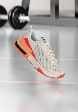 Beige en oranje sportieve sneaker met mesh bovenwerk en witte veters, zwevend boven een gepolijste sportschoolvloer met wazige fitnessapparatuur op de achtergrond.