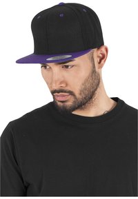 Cappellino da baseball nero con visiera piatta viola, dotato di occhielli viola e una toppa grigia sul davanti. Indossato da una persona in una maglietta nera.