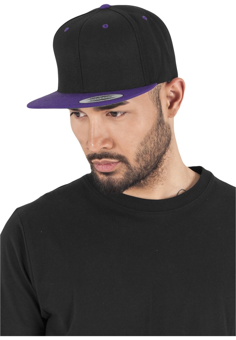 Cappellino da baseball nero con visiera piatta viola, dotato di occhielli viola e una toppa grigia sul davanti. Indossato da una persona in una maglietta nera.