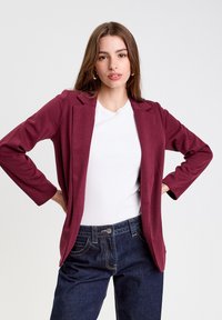 Blazer burdeos con cuello de solapa, mangas largas y diseño de frente abierto; combinado con una blusa blanca y jeans de mezclilla oscuros. Tejido texturizado, sin cierres.