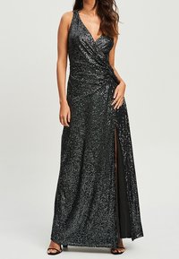 Femme portant une robe portefeuille noire sans manches ornée de sequins, avec une fente haute sur la cuisse, et des talons ouverts noirs, sur un fond neutre.