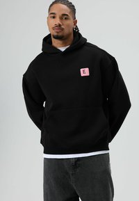 Zwarte hoodie van katoen, met een voorzak en een roze vierkante patch met de letter "E" op de linkerborst. Grijze spijkerbroek maakt de outfit compleet.