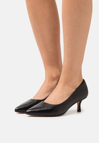 Chaussures à talons hauts en cuir noir avec un nez pointu et un petit talon. Texture lisse et design minimaliste, avec une découpe ouverte.