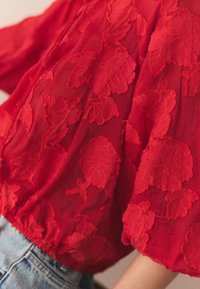 Červená priehľadná blúzka s kvetinovou výšivkou, krátkymi naberanými rukávmi, zbieraným lemom a jemnou textúrou. Kombinovaná so svetlomodrými denimovými nohavicami.