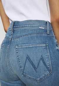 Jeans de mezclilla de lavado medio con cintura alta, que cuentan con bolsillos traseros y costuras azules en patrón de zigzag. Etiqueta visible en la cinturilla.