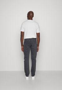 Vaqueros grises de corte ajustado con bolsillos traseros, combinados con una camiseta de manga corta blanca. Modelo de espaldas sobre un fondo neutro.