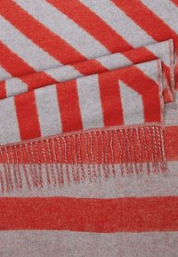 Écharpe en laine pliée avec des rayures diagonales et horizontales alternées rouges et gris clair, présentant des bords à franges rouges et grises.