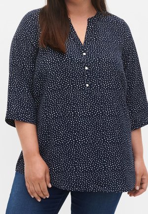Frau trägt marineblaue Tunika mit weißen Polka-Dots und dreiviertel Ärmeln, kombiniert mit dunkelblauen Jeans, steht vor einem einfarbigen Hintergrund.