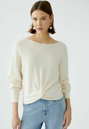 Light beige ribbed knit sweater with wide boat neckline, long sleeves, and twisted front detail, paired with light blue denim jeans.