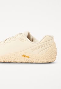 Light beige sneaker with textured rubber sole, featuring a wavy design and the "Merrell" logo embossed on the side. Highlights include a Vibram logo.