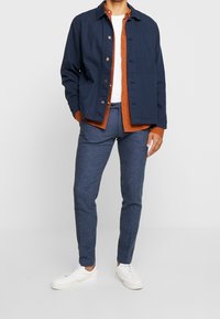Navy blue jacket with button details over an orange shirt, worn with textured blue trousers and white sneakers.