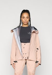 Casaco rosa à prova d'água com detalhes em cinza, apresentando capuz, fecho de correr e punhos ajustáveis. Usado sobre uma blusa cinza escura e calças combinando.