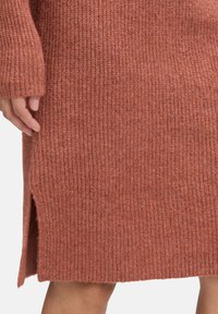 Close-up van een hand naast een jurk van ribbelbrei in roestkleur, met een lengte tot de knie en een zijsplit.