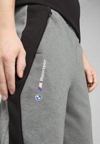 Sive hlače od somota s crnim detaljima, sa teksturiranom površinom, bočnim džepovima i "Motorsport" brendiranjem s BMW logotipom na boku.