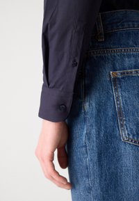 Navy button-up shirt with long sleeves, featuring a smooth texture and a single button cuff, paired with faded blue denim jeans.