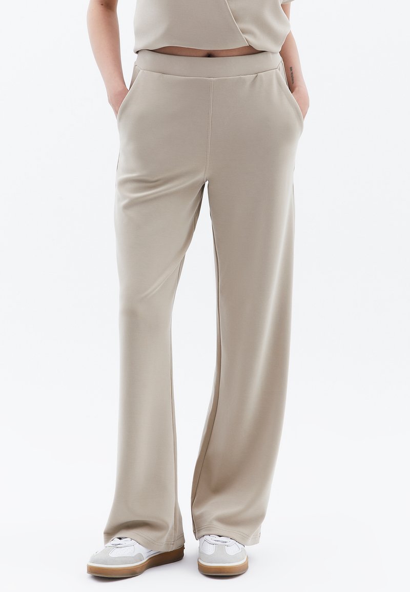 Beige, zachte stof, wijde broek met zijzakken, elastische tailleband en een gladde textuur, gecombineerd met witte sneakers op een effen achtergrond.
