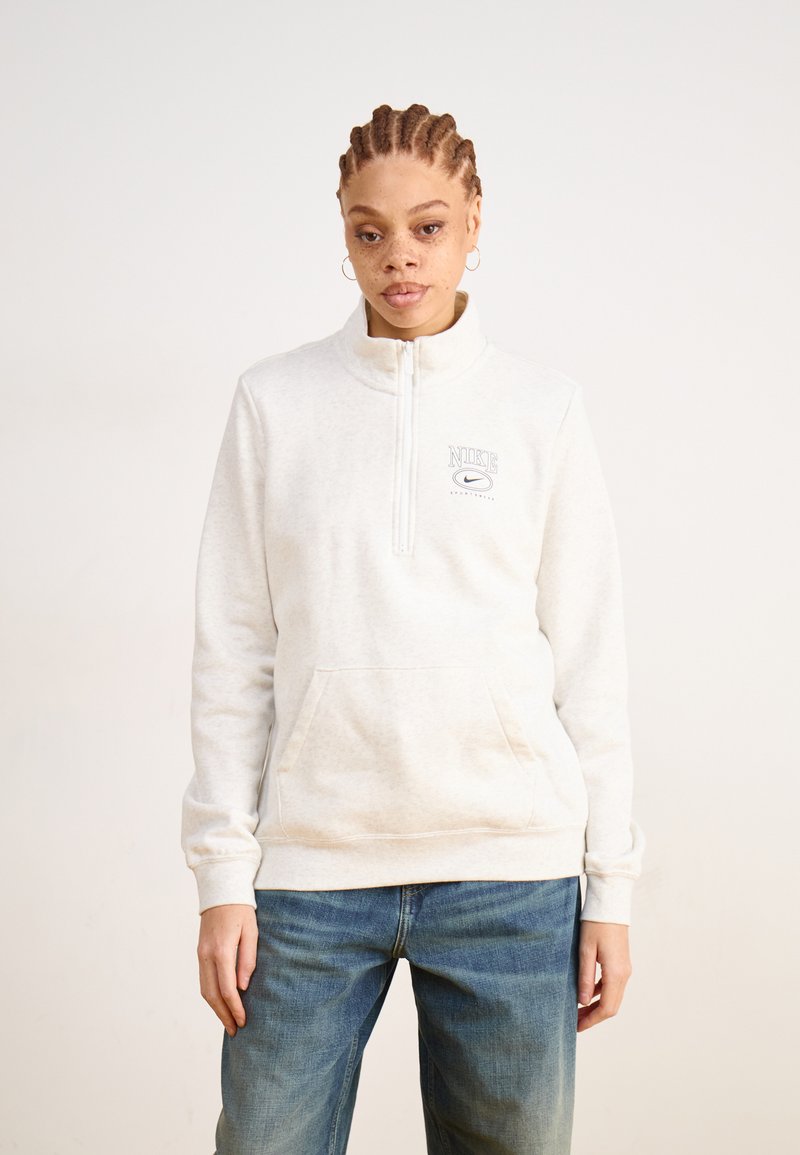 Lysegrå hettejakke med glidelås, høy krage, frontlomme og logoaccent. Mykt stoff og løs passform, kombinert med blå jeans.