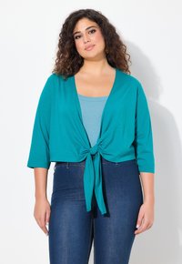 Cardigan court teal avec des manches trois-quarts, noué à l'avant ; superposé sur un haut bleu clair ; associé à un jeans en denim sombre.